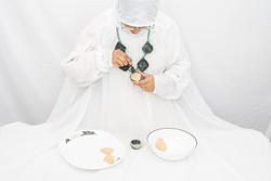 A person draped in white wearing a green necklace with gray accent pieces is kneeling in front of a plate and bowl.