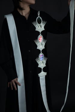 A student in black is wearing a long jewelry piece with white ribbon and ornate white pieces.