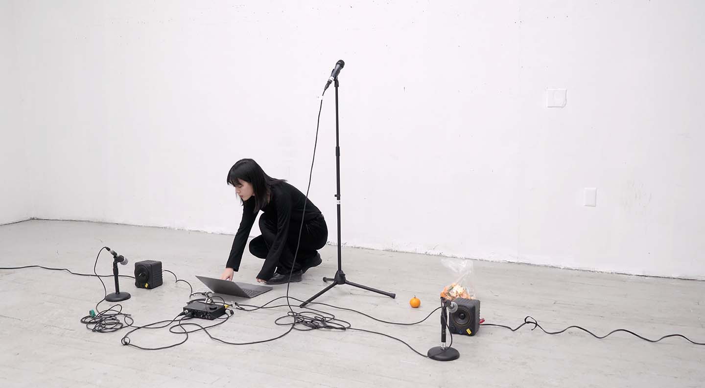 Image of student dressed in black performing in a white studio space with a laptop, three microphones, two speakers. An orange sits next to the student.